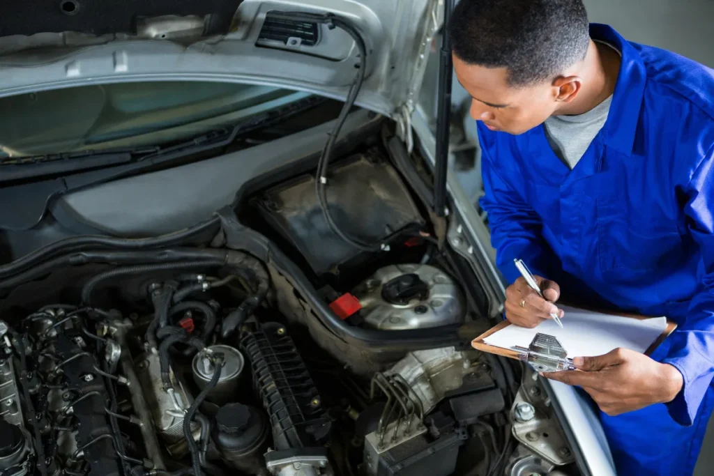 Auto mechanic in blue uniform inspecting car engine and writing diagnostic report on clipboard