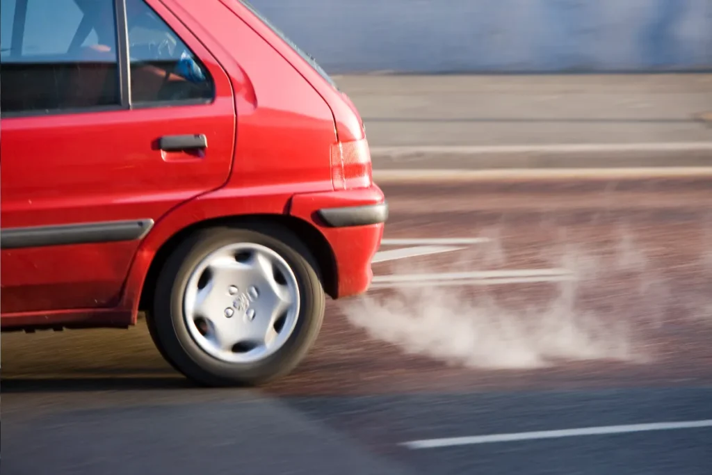 Red car accelerating with visible exhaust smoke, indicating possible engine or vacuum leak issue on the road